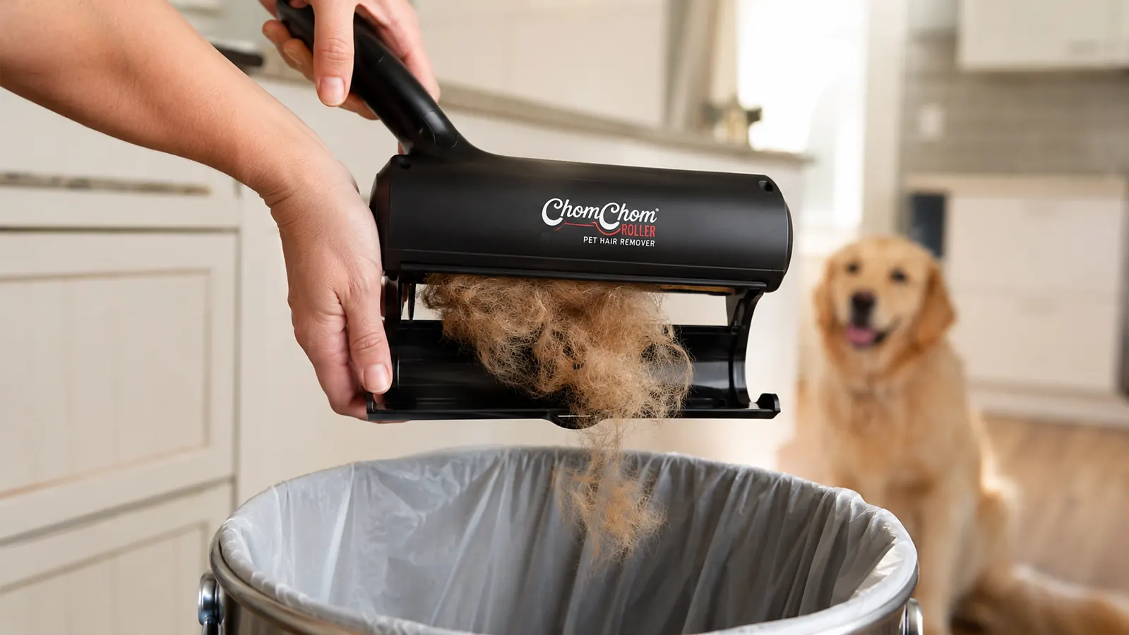 A hand in a kitchen holding the black ChomChom Roller open and tipping gathered dog hair into an open trash can, light brown fur wisping out of the chamber