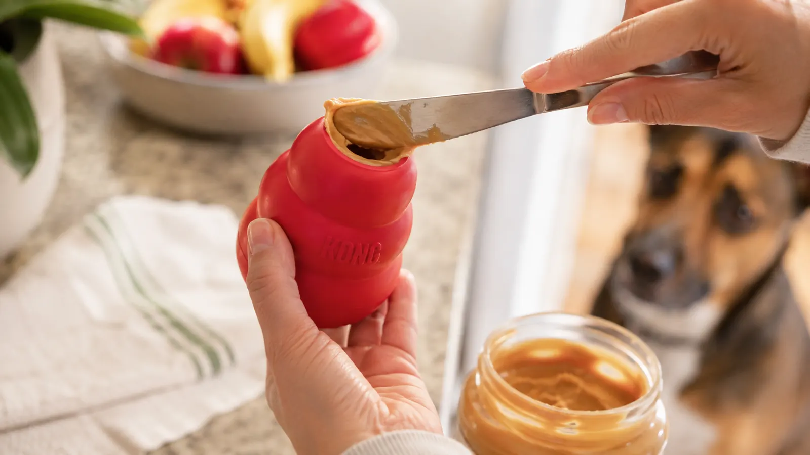 A hand spreading peanut butter into the hollow of a red KONG toy with a butter knife at a kitchen counter