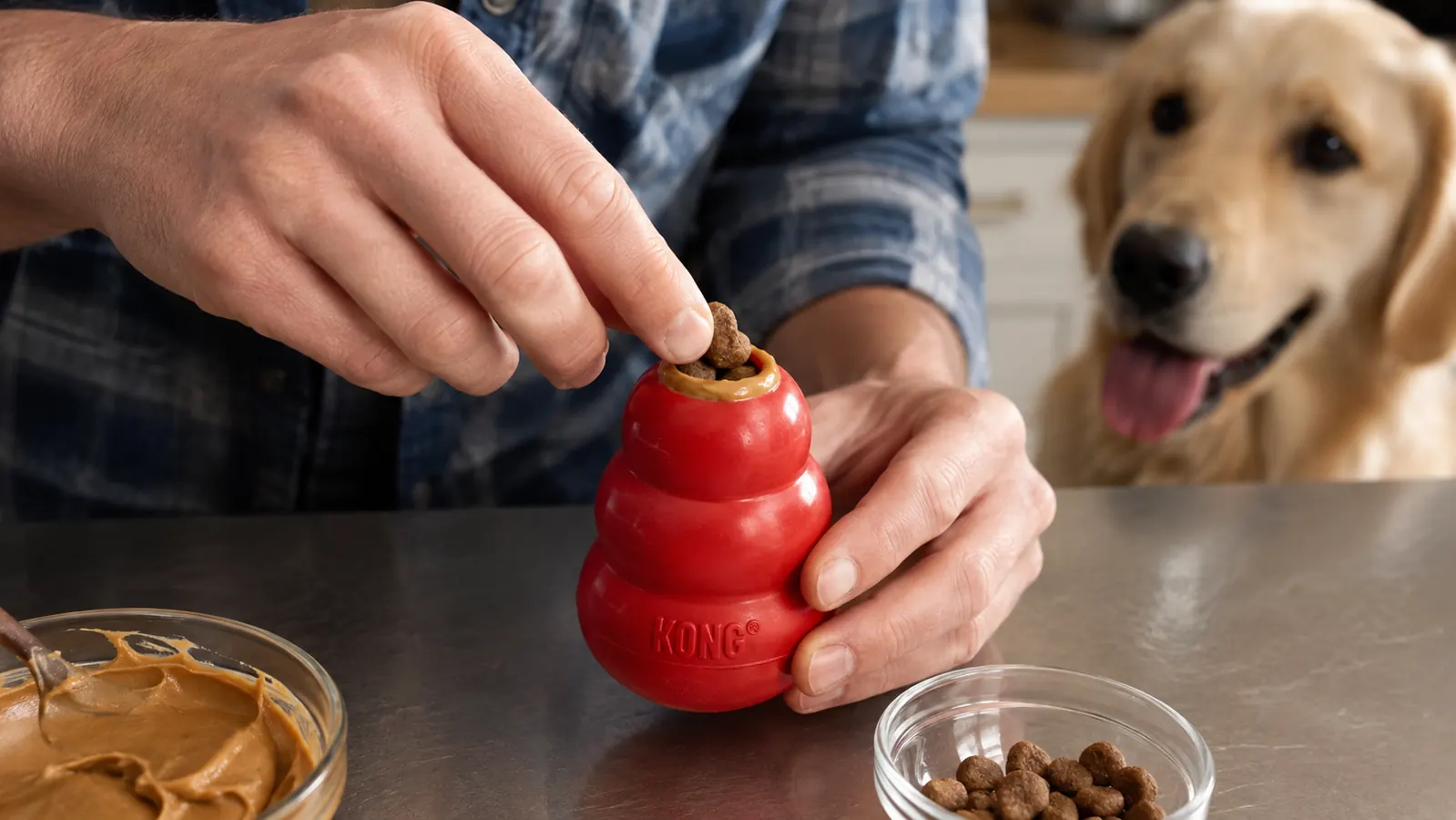 Hands packing a KONG Classic with peanut butter and a few bite-size treats, the smaller top opening filled first
