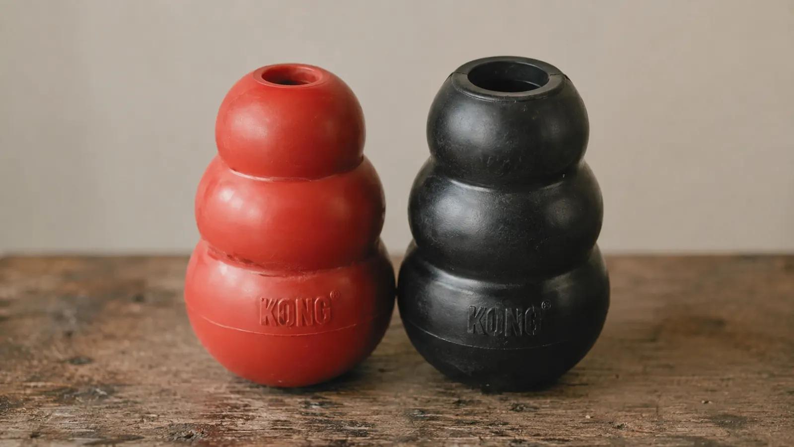 Red KONG Classic side by side with its black Extreme version, the black one visibly thicker walled, on a wooden table