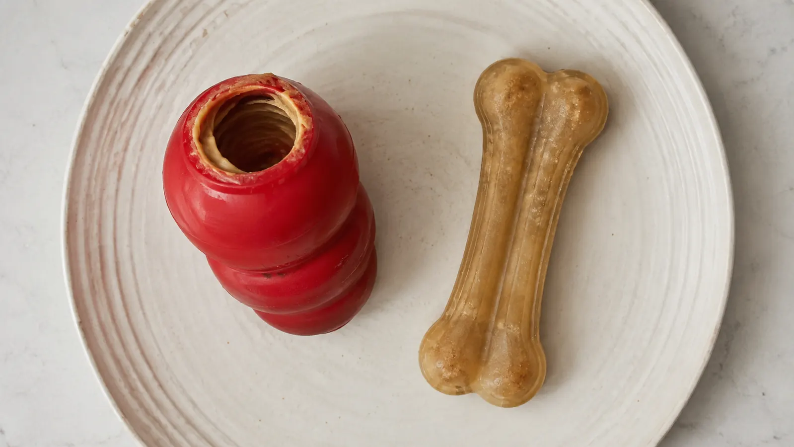 A top-down view of a red rubber KONG Classic toy sitting open on a small plate with peanut butter smeared inside, next to a plain bone-shaped flavored chew